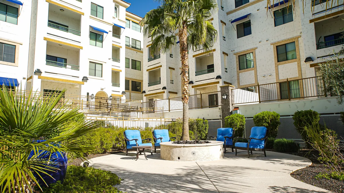 This exterior image showcases a welcoming outdoor space with a modern fire pit surrounded by blue lounge chairs, a tall palm tree, and lush greenery. The arrangement of furniture invites both small gatherings and individual relaxation, reflecting a cozy and social environment.
