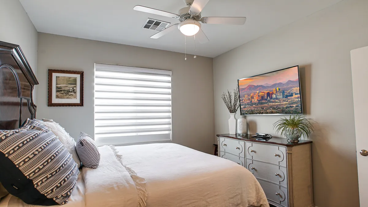 A cozy guest bedroom with a king-size bed dressed in decorative pillows, a dresser topped with a TV, a window with blinds letting in natural light, and framed wall art, offering comfort and ample storage for guests.