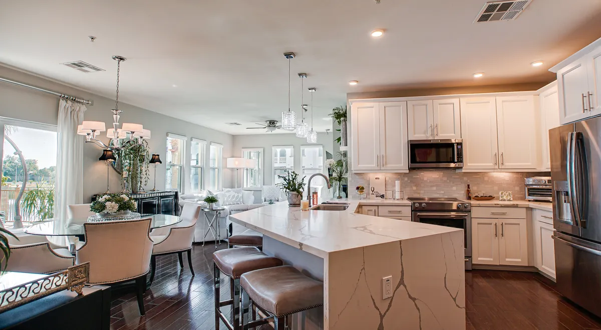 This image showcases a cohesive open-plan living space that seamlessly blends the kitchen island, dining area, and living room seating. Large windows flood the room with natural light, and the overall aesthetic is both inviting and stylish. The space is clearly designed for both relaxing and entertaining, corresponding to the heart-of-the-home atmosphere described in the section.