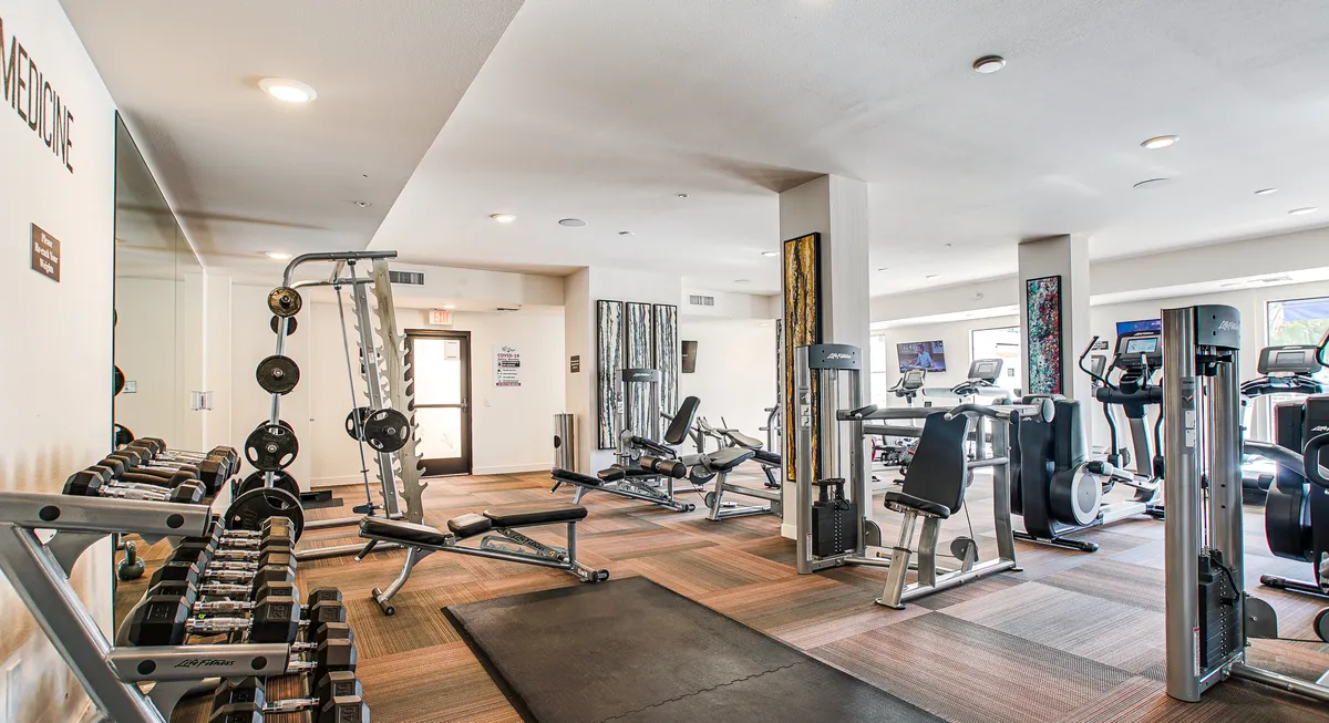 A fitness center interior with a rack of dumbbells, state-of-the-art exercise machines, mirrored walls, and exercise mats. The atmosphere is energetic, motivating residents to pursue wellness goals from the convenience of home.