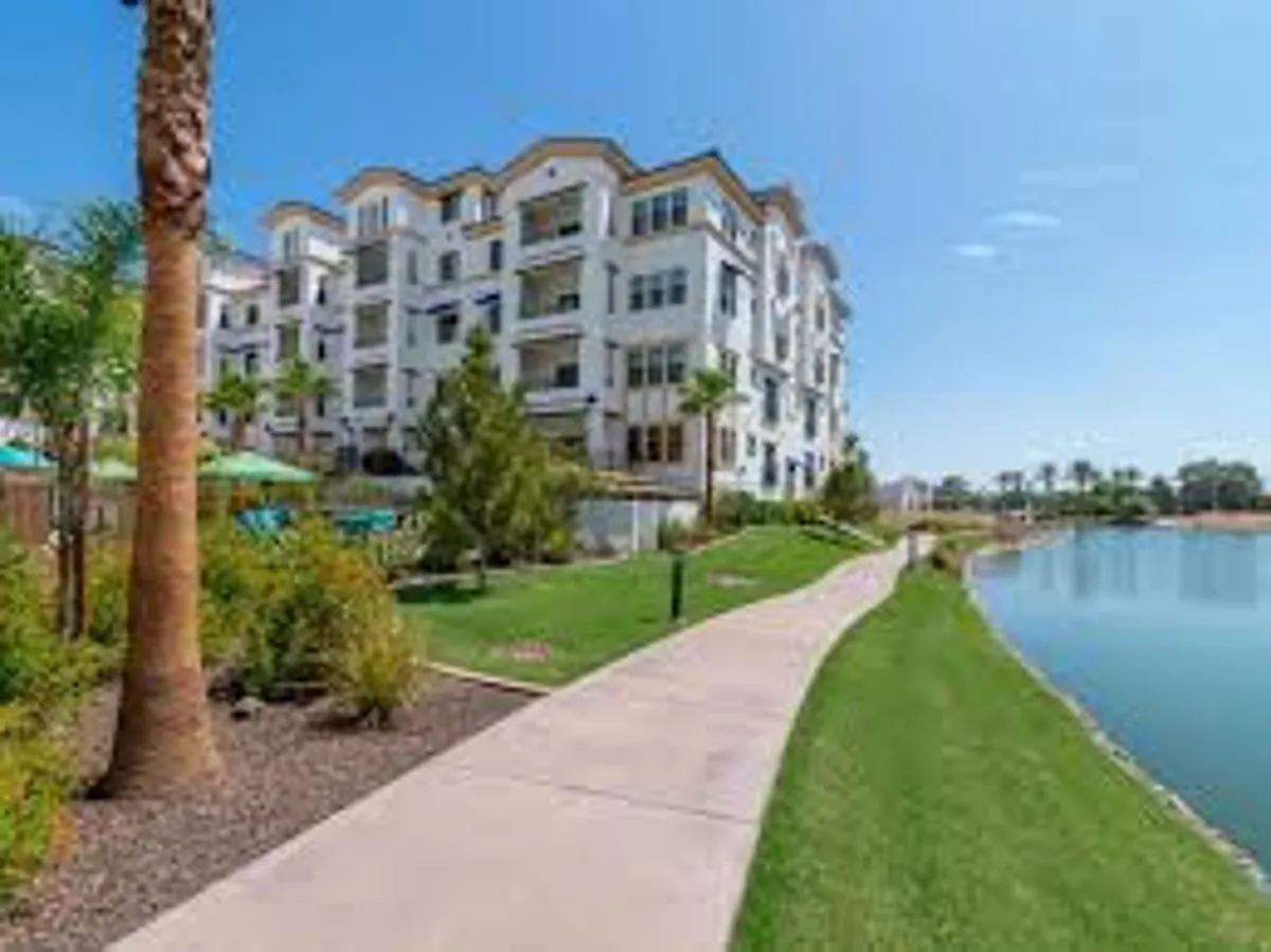 A tranquil exterior scene featuring a modern apartment building situated beside a serene lake. The landscaped pathway invites residents and visitors to stroll along the water, with lush greenery and open space highlighting the natural beauty of the area.