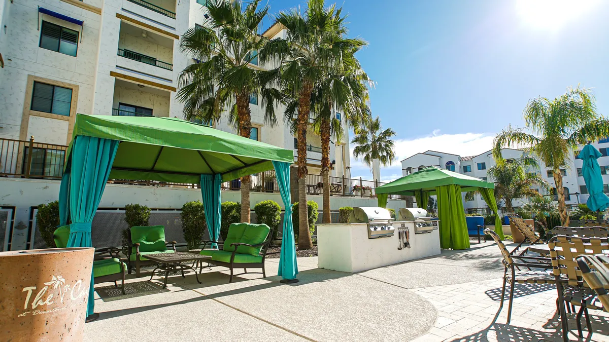 An outdoor seating area shaded by green canopies and palm trees, featuring a barbecue grill and plenty of space for social gatherings. The relaxed, cheerful mood suggests ideal settings for barbecues, lounging, and friendly get-togethers.