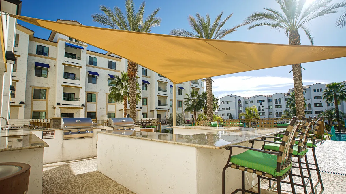 An inviting outdoor space featuring a modern outdoor kitchen area, barbecue grills, ample seating with plush green cushions, shade sails, palm trees, and a glimpse of the pool in the background. The setting is designed for social gatherings, dining, and poolside relaxation.