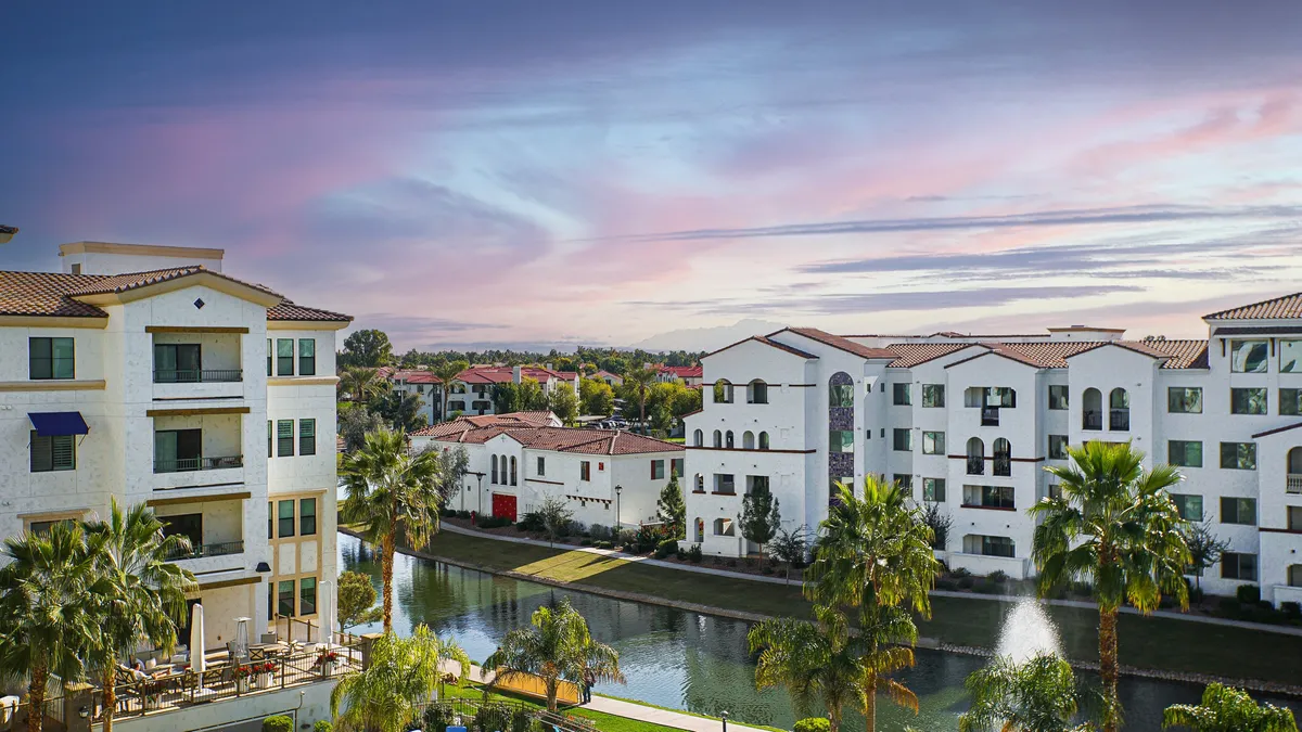 This image features modern architecture surrounded by palm trees and a serene water canal, with colorful rooftops and people enjoying the outdoor spaces. The scene conveys the tranquility and upscale environment of the community, highlighting the resort-like setting and proximity to water features reminiscent of a golf course landscape.