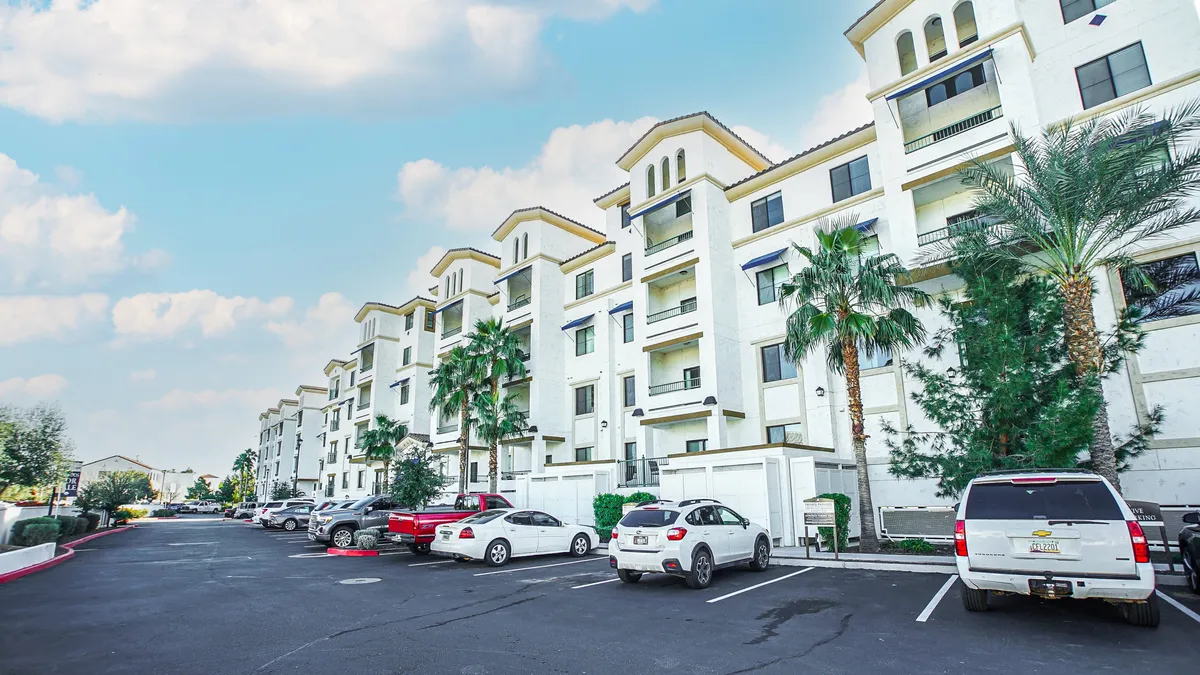 A welcoming exterior view of modern apartment buildings surrounded by palm trees and a parking lot. The scene feels inviting and suggests easy access to both residential and local amenities, with sunny weather and a sense of community.