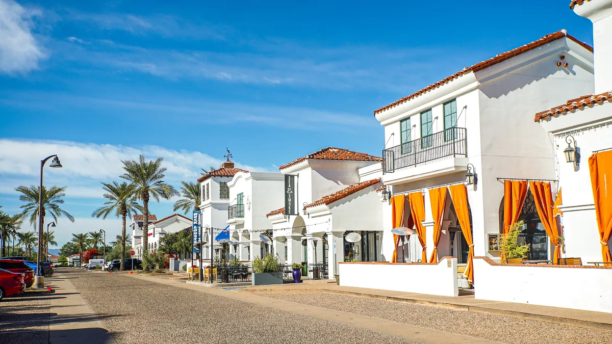 A lively exterior view of an outdoor dining area, framed by colorful awnings, palm trees, and distinct architectural details. People are seen dining and socializing, suggesting an active, vibrant community life adjacent to the home environment.