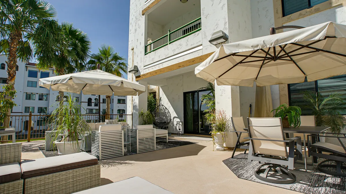 A serene patio setting is displayed with multiple outdoor seating options including a hanging chair, umbrellas, planters, and palm trees. The area looks perfect for unwinding, socializing, or enjoying a meal outdoors, with soft sunlight casting a calming glow.