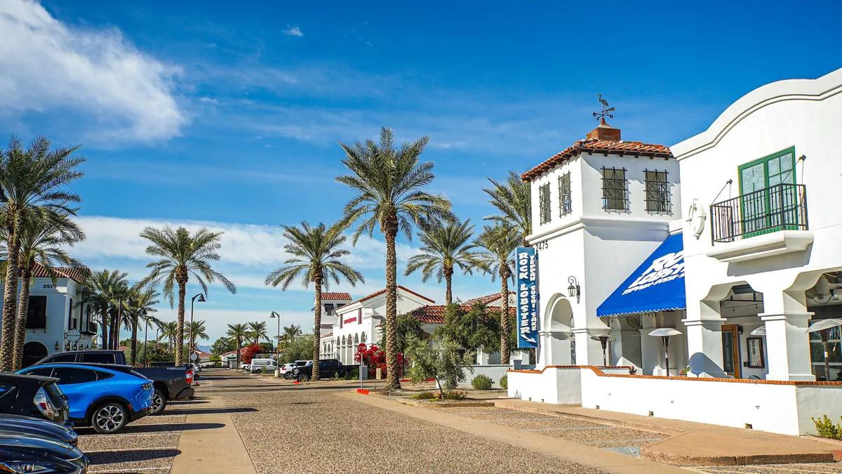 This image features a palm-lined, cobblestone street with white stucco buildings and a blue awning. The scene captures a relaxed atmosphere where people might stroll, shop at local boutiques, or enjoy outdoor dining. The overall mood is vibrant and inviting, echoing the ambiance of a lively downtown area.