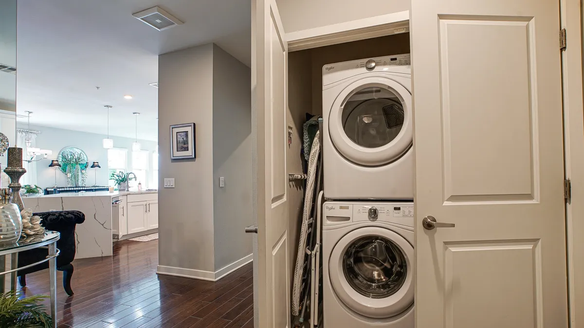 A practical and efficient laundry room is depicted with a stacked washer and dryer, a doorway leading toward the kitchen, and a decorative plant on the table. The space appears tidy and functional, reinforcing the promise of convenience for extended stays.